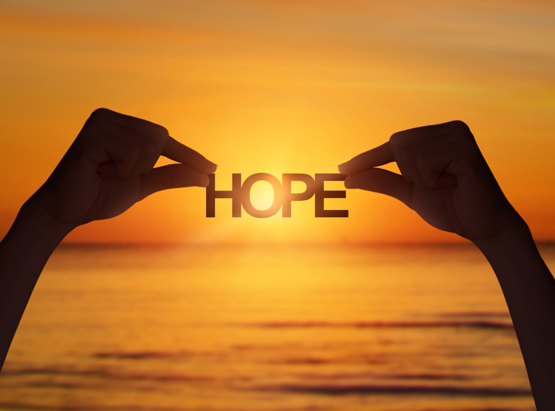 Photograph of the word ‘Hope’ held up with two hands, silhouetted against a orange sunny sky.